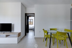 Un salon moderne avec un téléviseur à écran plat posé sur un meuble en bois, une table à manger en verre entourée de chaises jaunes, et une vue sur une chambre  à coucher à travers une porte ouverte.