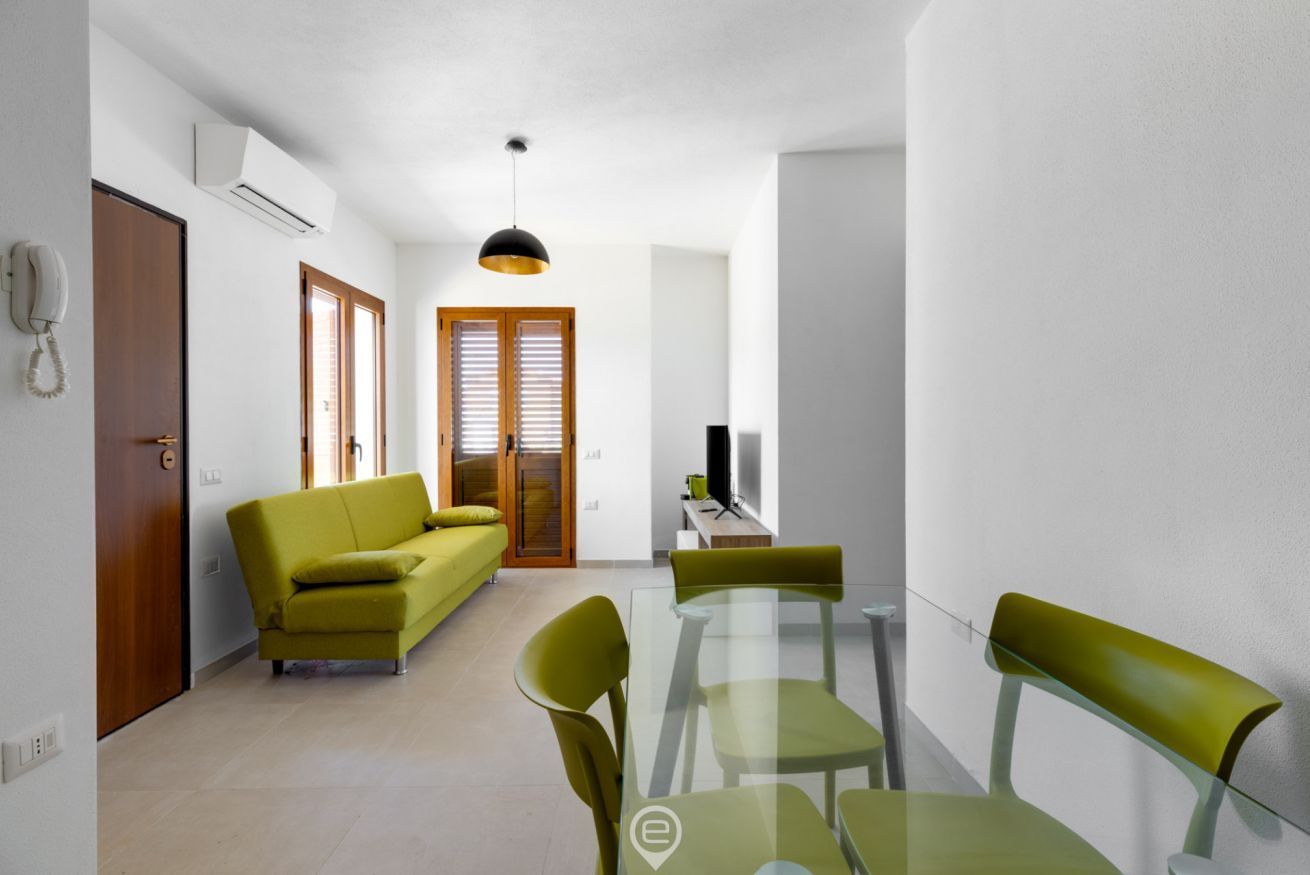 A modern living room with a sofa, a glass dining table, and chairs. There is a TV on a stand, a hanging light fixture, and a door with a phone on the wall.