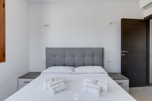 A modern bedroom with a grey tufted headboard, white bedding, and neatly folded towels on the bed.