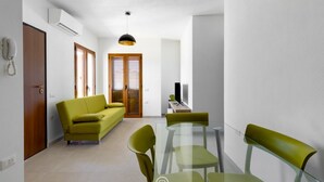 Uma sala de estar moderna com um sofá, uma mesa de jantar de vidro e cadeiras. Há uma TV sobre um suporte, uma luminária pendente e uma porta com um telefone na parede.