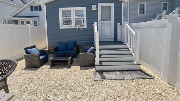 A small, single-story building with a gray exterior, a front door, and a staircase leading to a porch. There are wicker chairs and a table on the porch.