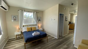 A bedroom with a bed, bedside tables, a window with curtains, a ceiling fan, and a hallway leading to a kitchen. - Retro Retreat - A Desert Oasis (Twentynine Palms)