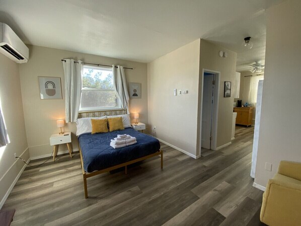 A bedroom with a bed, bedside tables, a window with curtains, a ceiling fan, and a hallway leading to a kitchen.