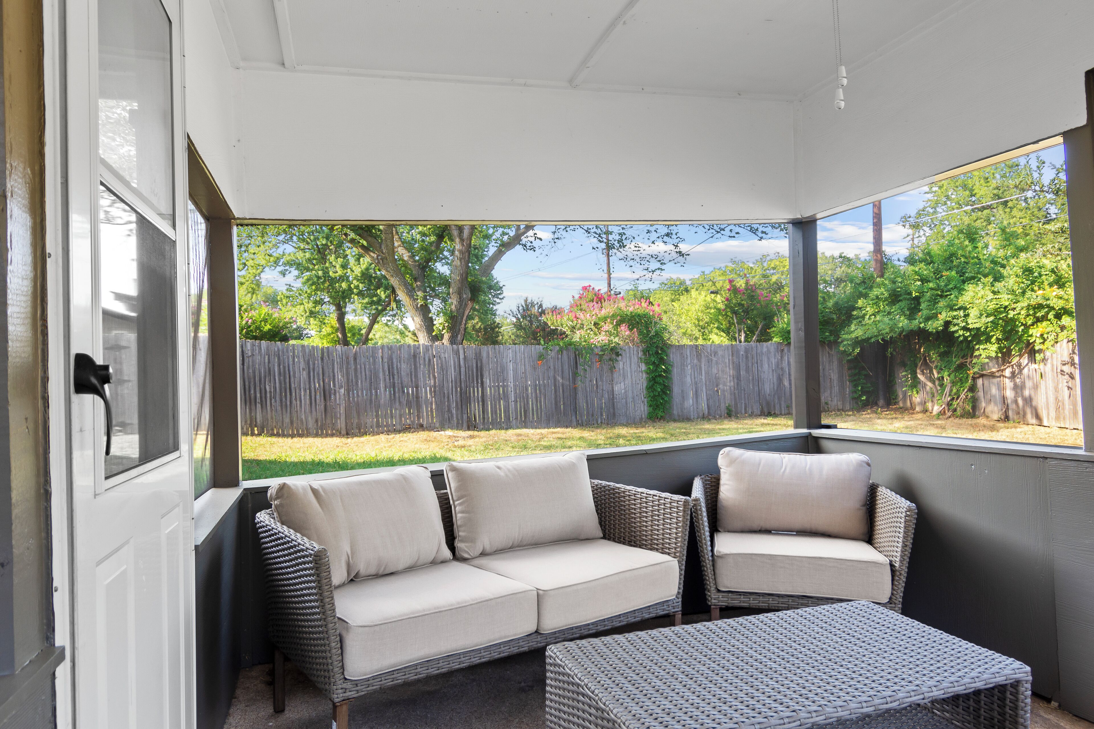 A covered patio area with a wicker sofa and chair, a small table, and a view of a backyard with a wooden fence and trees.