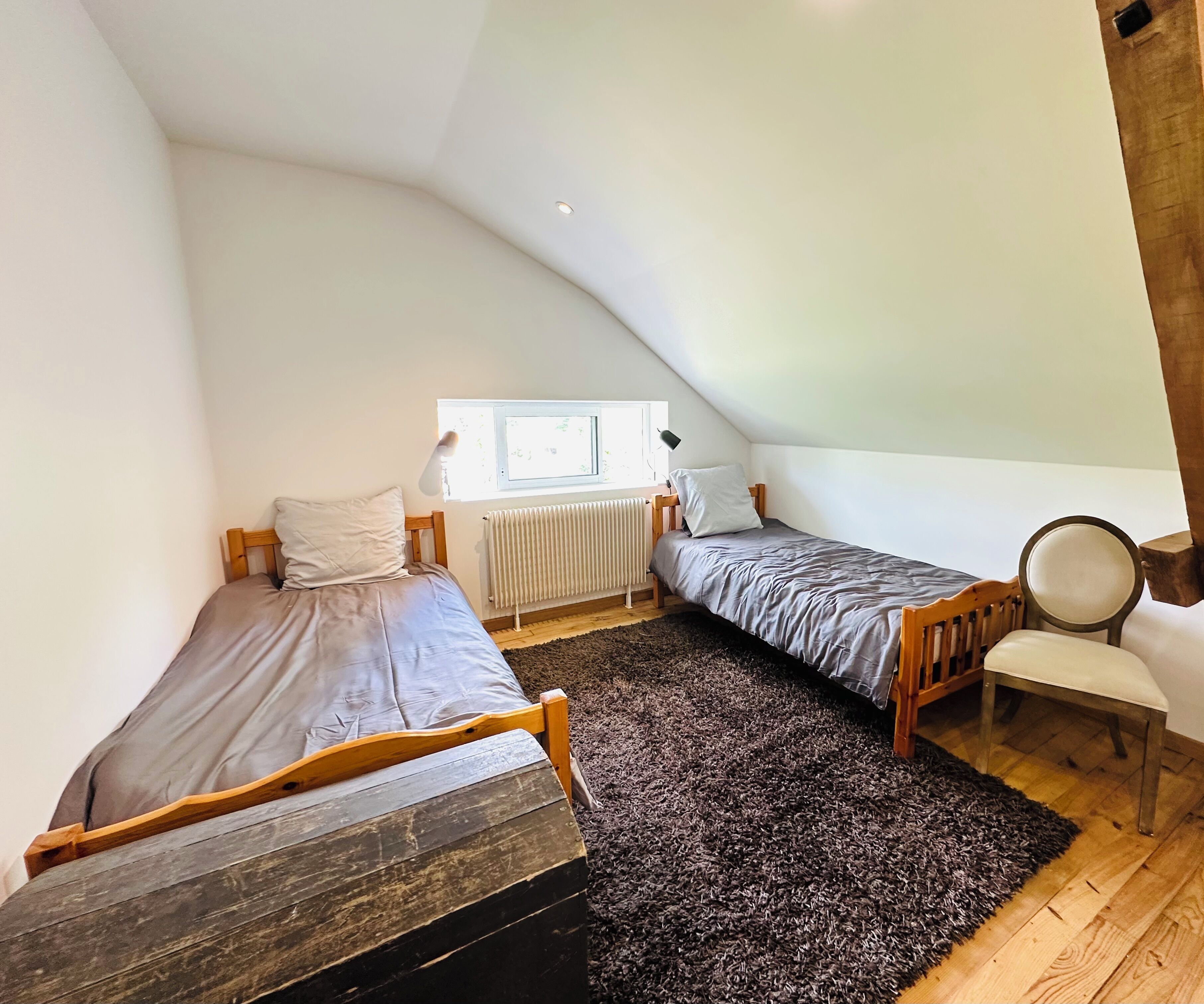 Une petite chambre mansardée, chaleureuse et accueillante, avec deux lits simples, un radiateur, une fenêtre et une chaise.