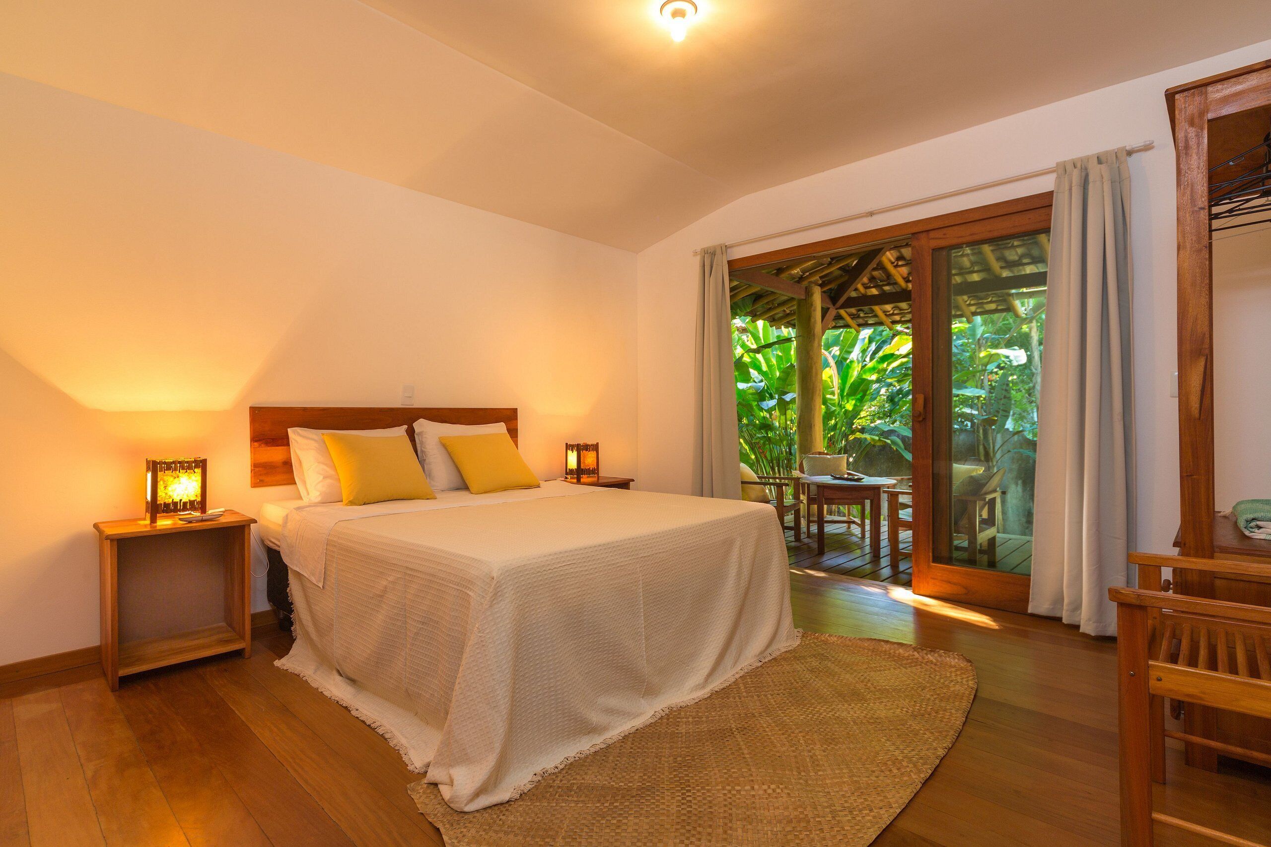 Un dormitorio con cama, mesitas de noche, una silla de madera y vistas a un jardín a través de un ventanal.