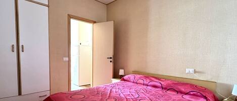 A bedroom with a bed, a wardrobe, a bedside table, and a door leading to another room.