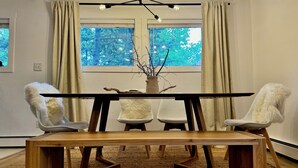 Ein Essbereich mit einem Holztisch, vier weißen Stühlen, einer Vase mit Zweigen und einem Fenster mit Vorhängen.