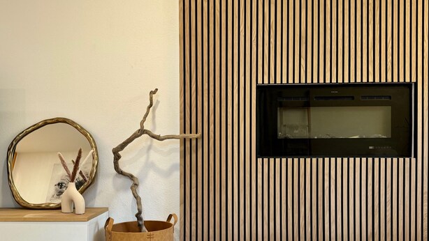 Ein modernes Wohnzimmer mit einer Wand aus Holzlamellen, einem Kamin, einem runden Spiegel und einer Vase mit Zweigen.