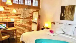 A bedroom with a brick wall, a desk with a computer, a bed with white bedding and a blue accent, and a painting on the wall. - Room #3 two blocks from the Malecon (La Paz)