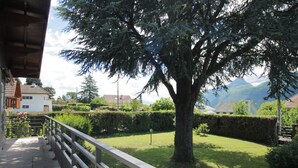 Un balcon doté d’une balustrade en métal, qui donne sur un jardin bien entretenu avec un grand arbre, une haie et une vue sur une chaîne de montagnes en arrière-plan.