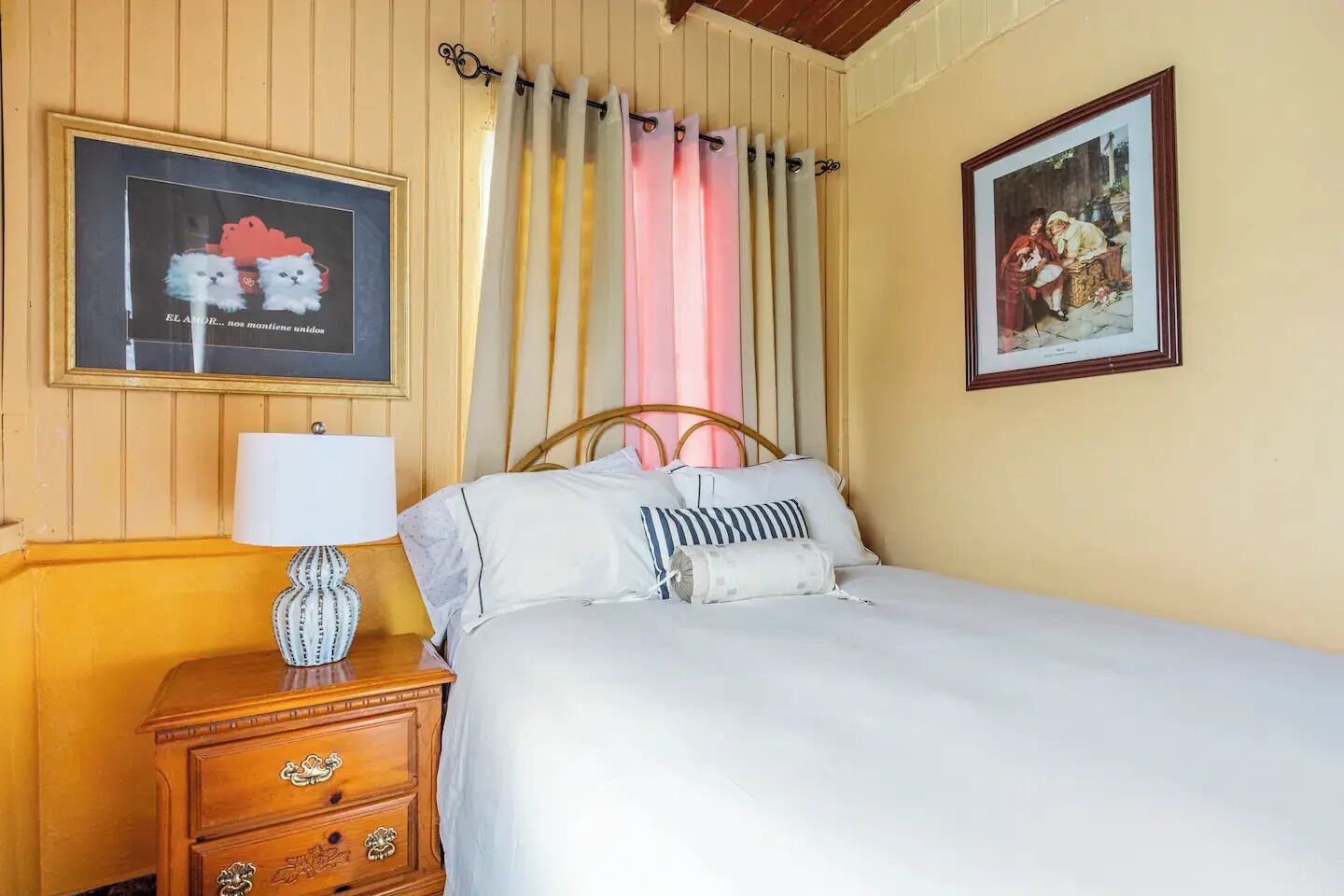 Un dormitorio con cama, mesita de noche con lámpara, cómoda de madera, un cuadro en la pared y una cortina.