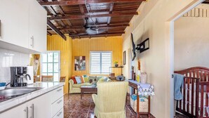 A kitchen with white cabinets and a coffee maker, a living area with a sofa and a chair, and a wooden ceiling.