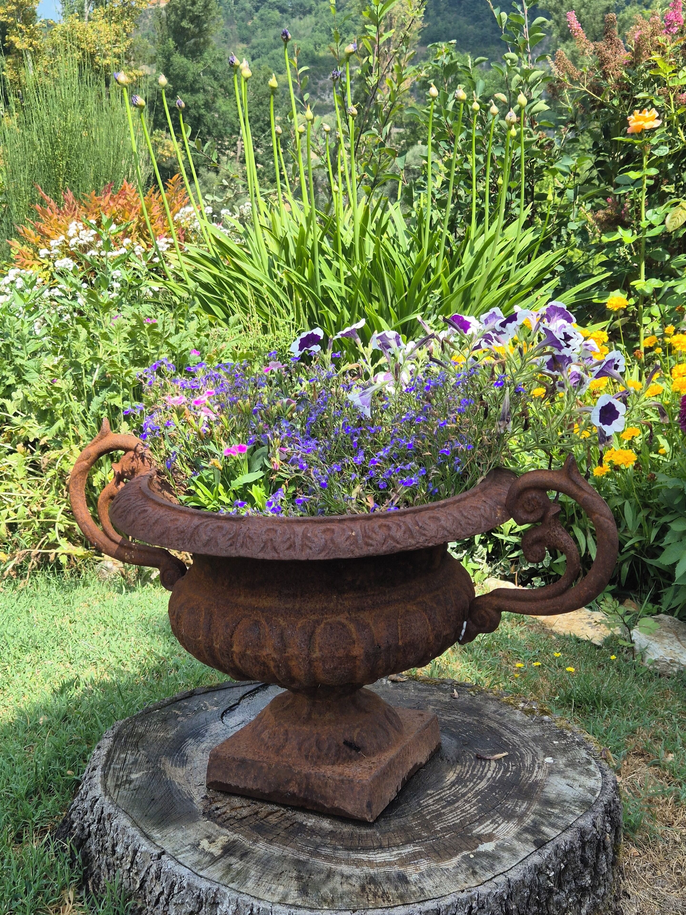 En rustik metalplanter med en træsokkel, fyldt med forskellige blomster og grønne planter, placeret i en have.