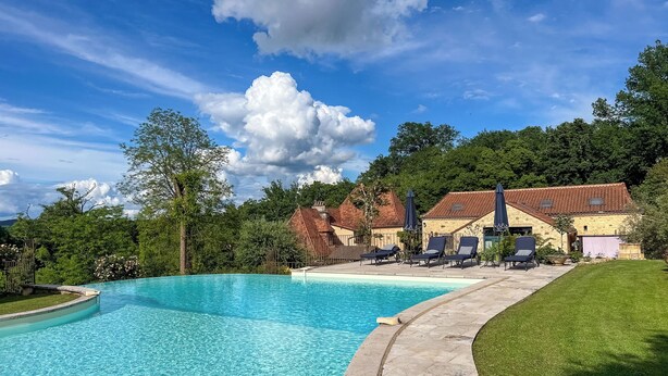 Ein Schwimmbecken mit Liegestühlen und Sonnenschirmen, umgeben von einem gepflegten Rasen und einem Gebäude mit einem Ziegeldach.