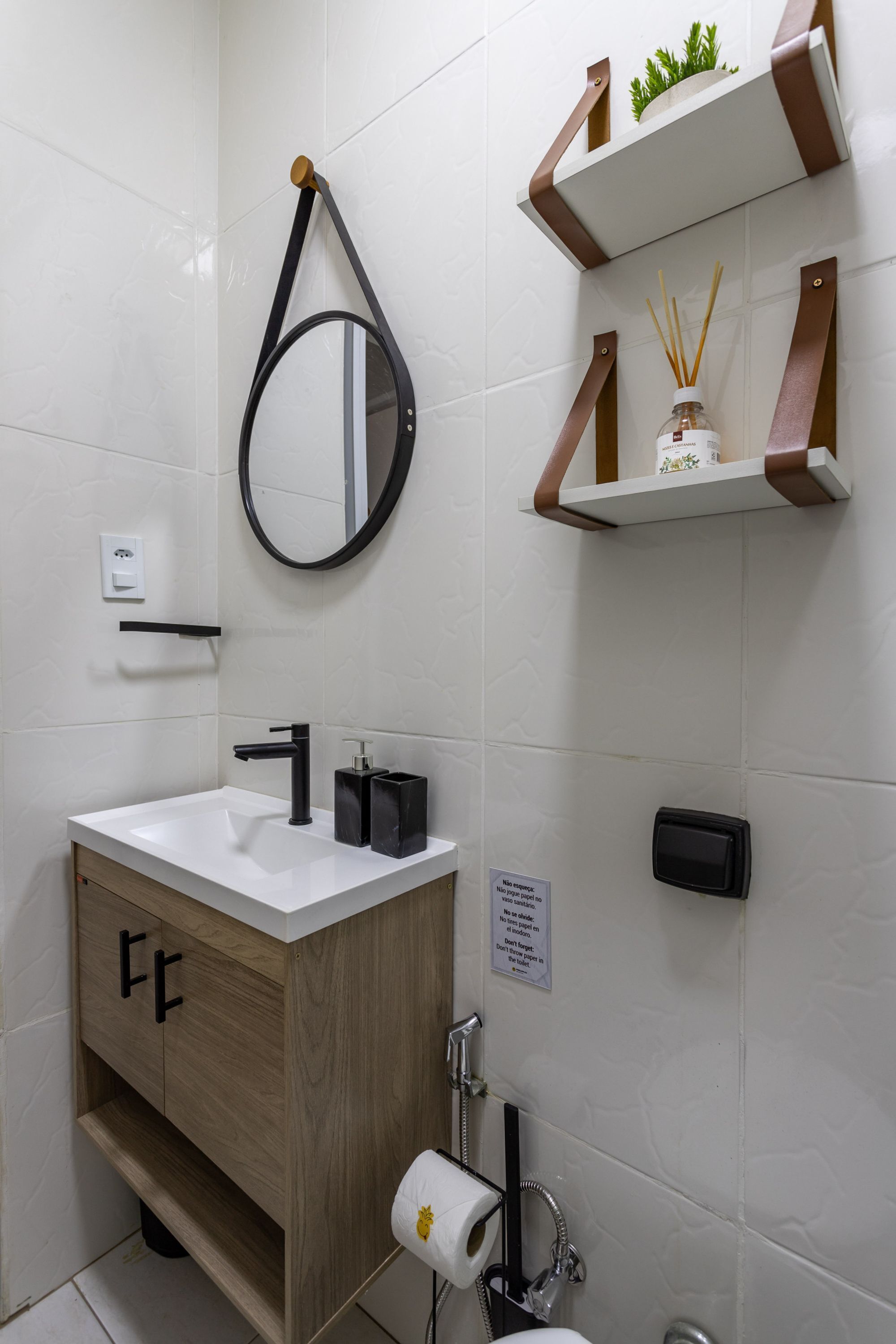 Un baño moderno con lavamanos blanco, grifo negro y mueble de madera. Hay un espejo con marco de madera, un dispensador de toallas y una estantería con una planta y difusor de aroma.