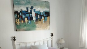 Een slaapkamer met een wit bed, een wit hoofdeinde, een nachtkastje met een lamp, een klein, wit nachtkastje en een abstract schilderij aan de muur.