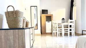 Ein modernes, helles Zimmer mit Schreibtisch aus Holz, weißen Stühlen, einem großen gewebten Korb und einem Flachbildfernseher auf einer Holzkommode.