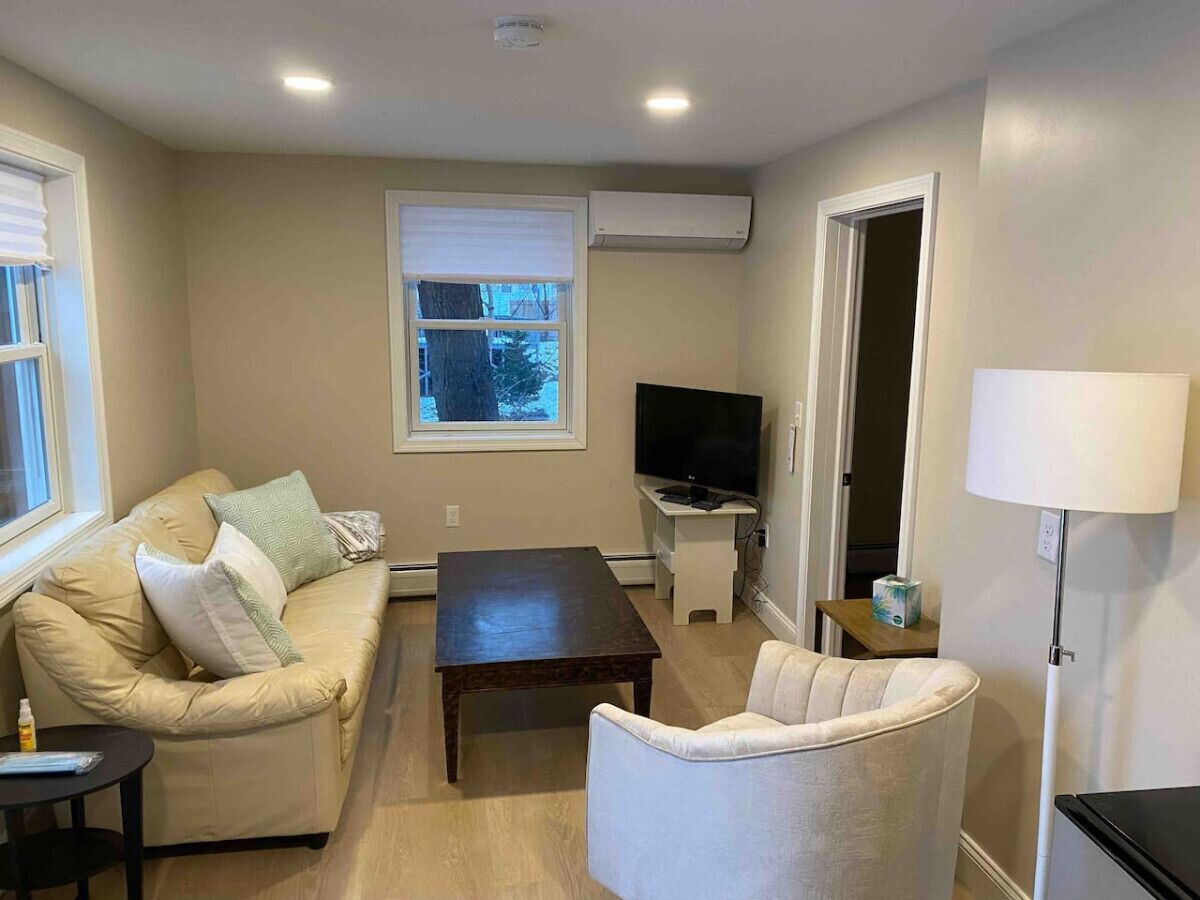 Een woonkamer met een beige bank, een witte fauteuil, een donker houten salontafel, een flatscreen televisie op een standaard en een vloerlamp.