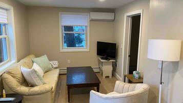 Een woonkamer met een beige bank, een witte fauteuil, een donker houten salontafel, een flatscreen televisie op een standaard en een vloerlamp.
