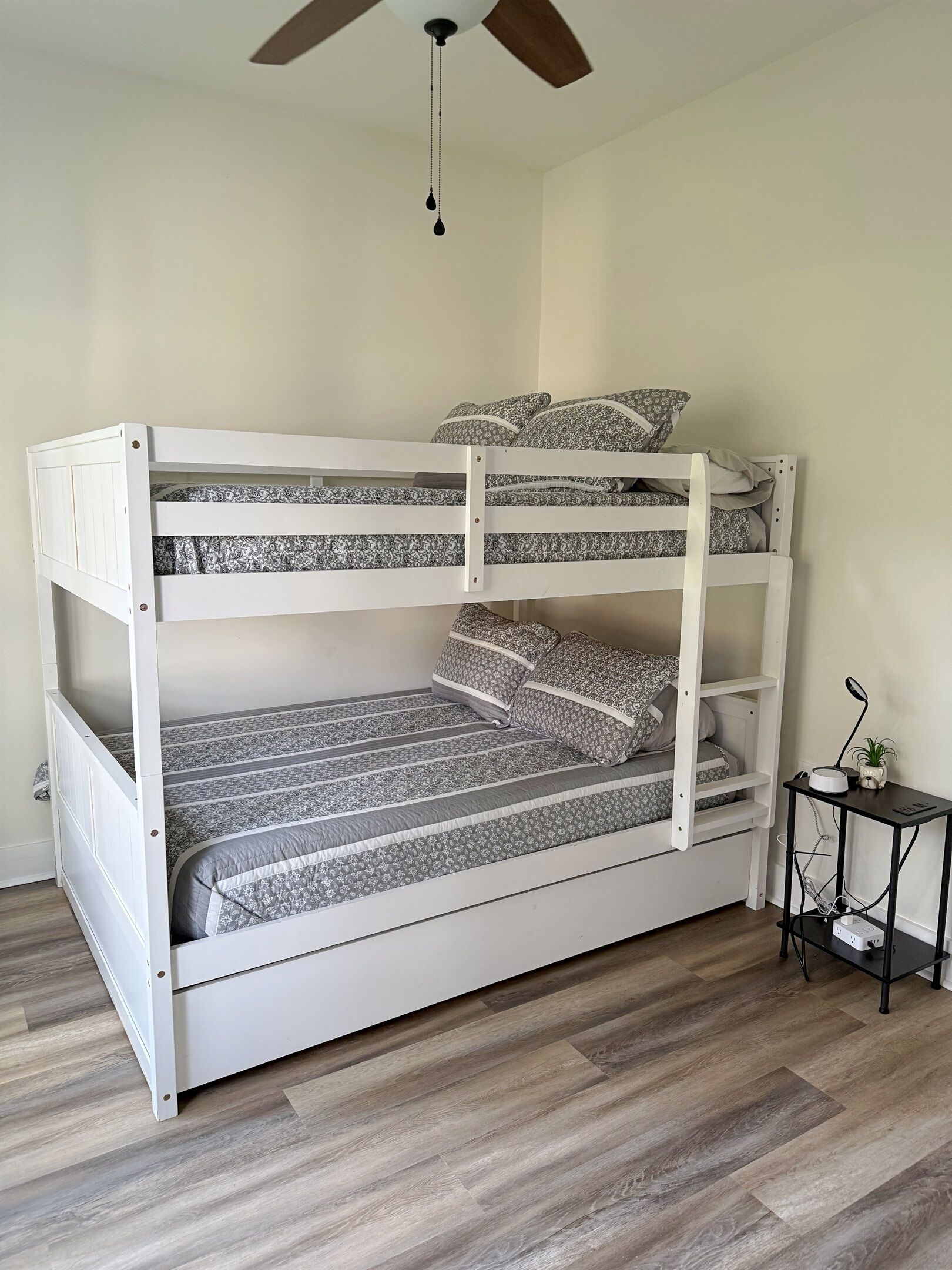 Un lit superposé avec une structure blanche, une literie grise et une petite table sur laquelle se trouvent une plante et une lampe.