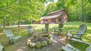 A cabin with a fire pit surrounded by Adirondack chairs, set in a wooded area. - Alden Home w/ Fire Pit: Steps to Torch Lake! (Alden)