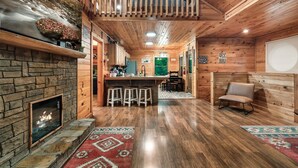 A cozy cabin interior with a stone fireplace, wooden floors, a kitchen area, and a balcony. - Stone-Built Cabin w/ Indoor Pool, Firepit, & Bunk Room @ Smokies Retreat! (Sevierville)