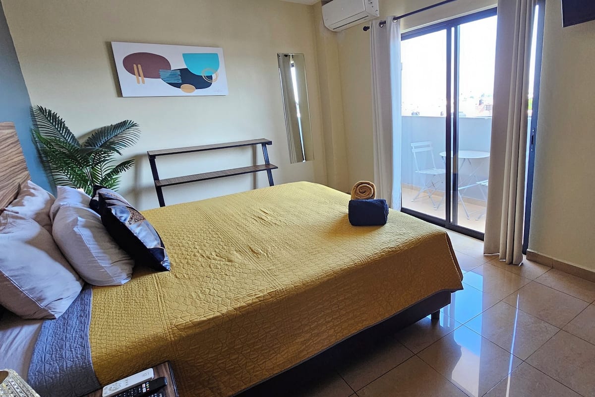 Quarto de hotel com cama, uma pequena mesa, um vaso de planta e vista para a varanda externa.