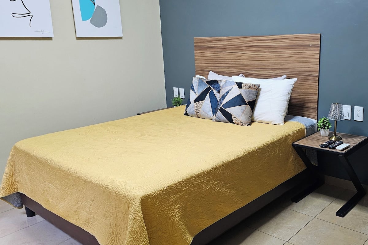 Uma cama bem arrumada com colcha amarela, cabeceira de madeira e almofadas decorativas. Há uma pequena mesa de cabeceira com um abajur e um controle remoto.
