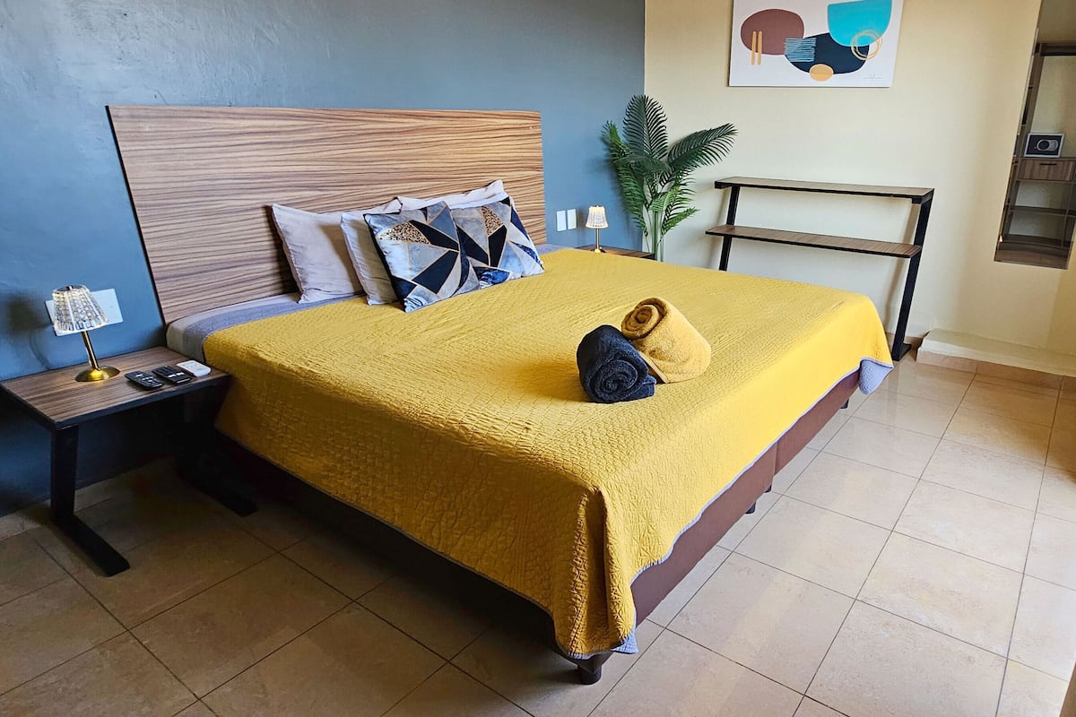 Cama bem arrumada com colcha amarela, cabeceira de madeira e almofadas decorativas. Tem uma mesa de cabeceira com abajur e uma pequena planta.
