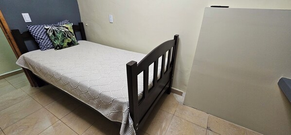 A single bed with a wooden frame and a textured white bedspread in a room with tiled flooring and a plain wall.