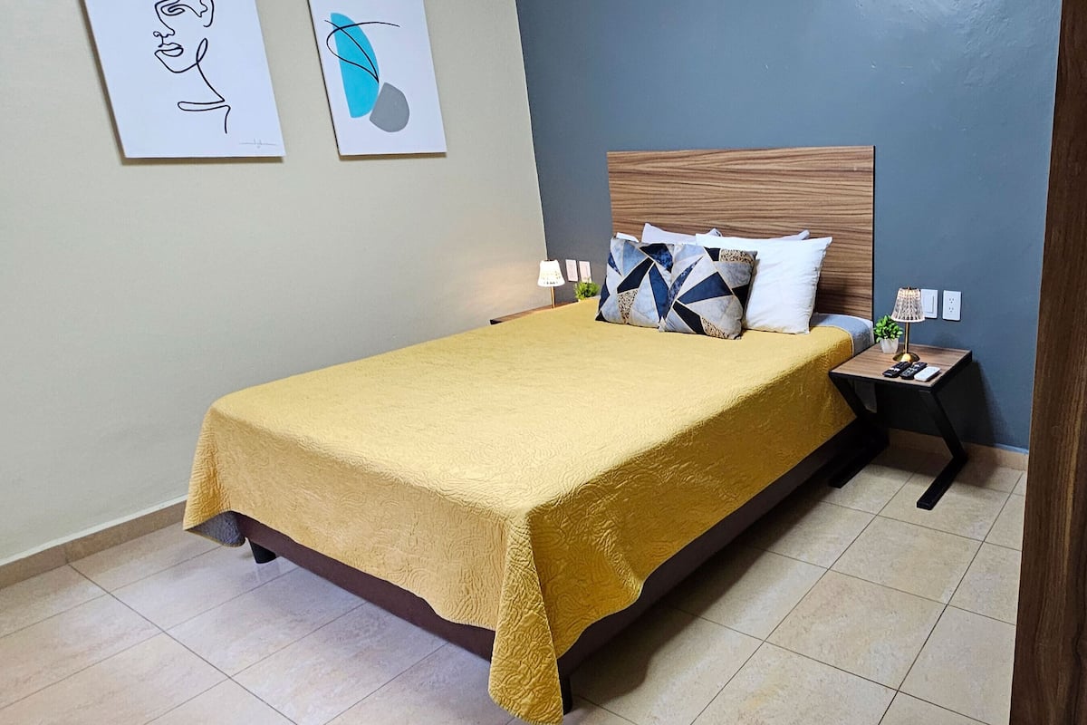 Uma cama bem arrumada com colcha amarela, cabeceira de madeira e duas luminárias de cabeceira.