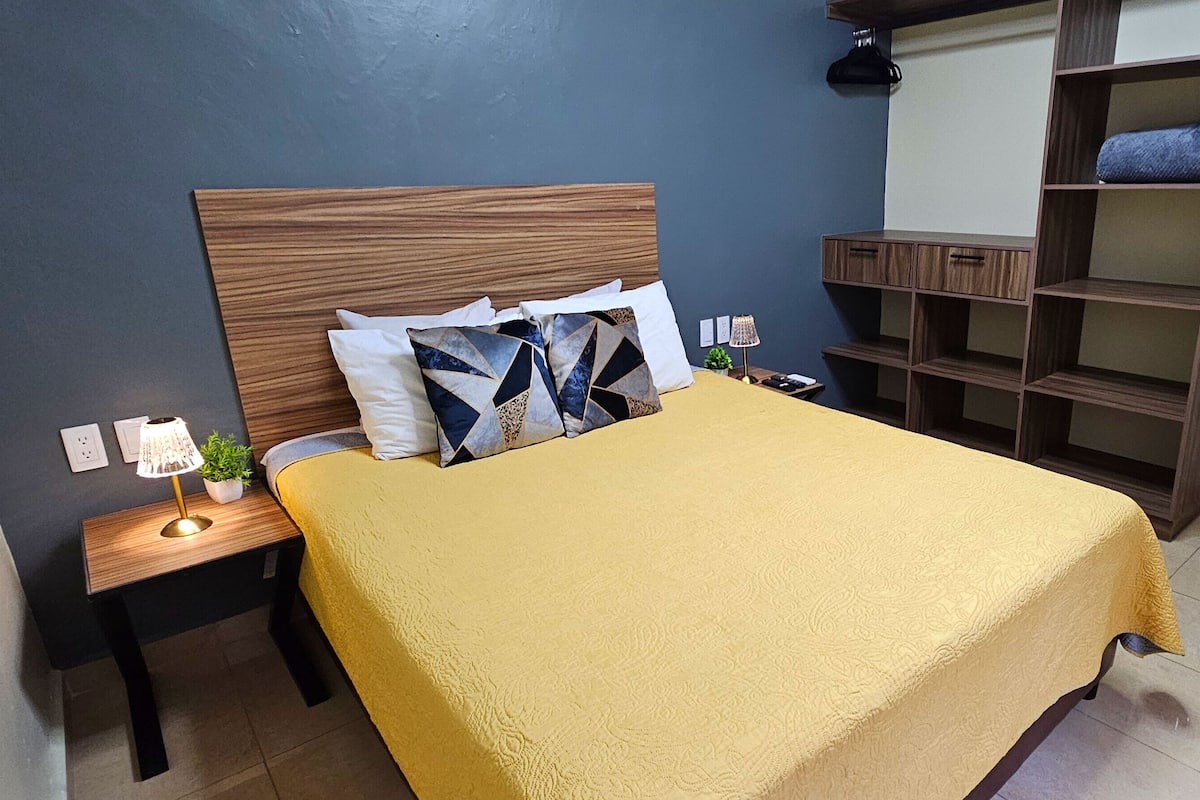 Quarto bem arrumado com cabeceira de madeira, colcha amarela e uma estante embutida na parede.