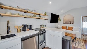 A modern kitchen with white cabinets, a built-in oven, and a dishwasher. There are wooden shelves above the counter holding kitchenware. A flat-screen TV is mounted on the wall, and a sofa with orange cushions is in the adjacent living area.