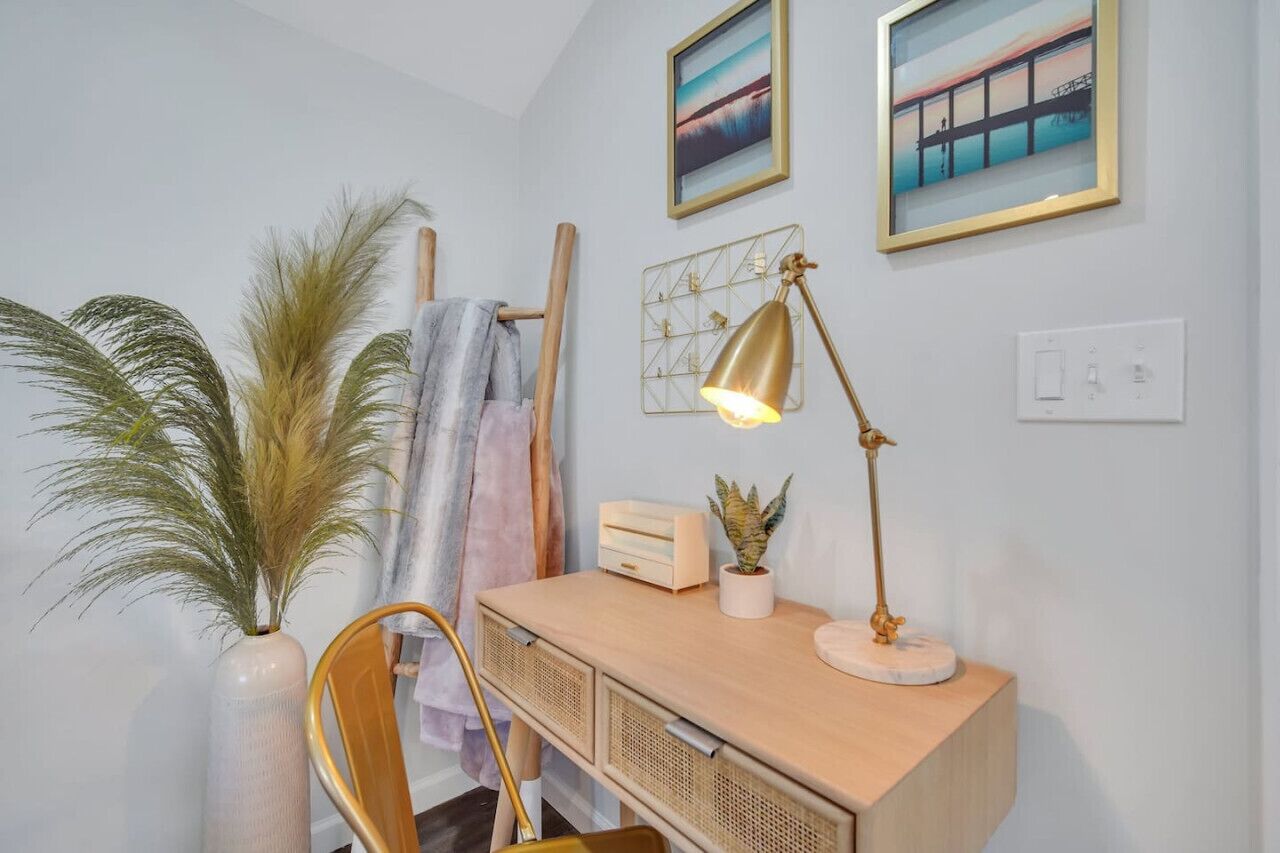 Una habitación con un escritorio de madera, una lámpara de escritorio dorada, una planta en maceta, una silla y dos cuadros en la pared.