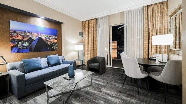 A modern hotel room with a cityscape view, a sofa, a dining table with chairs, and a large window with curtains. - PH Floor Studio • Strip View • Balcony • No Resort Fee D30O3 (Las Vegas)