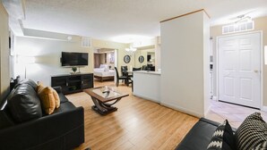 Un soggiorno moderno con un divano in pelle nera, un tavolino in legno e un televisore a schermo piatto. Si intravedono in secondo piano una camera da letto con un letto e un bagno con il water e il lavandino.