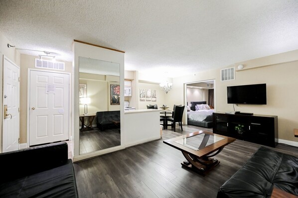 A modern living room with a large mirror, a flat-screen TV, a dining area, and a bedroom with a bed and nightstand.