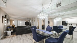 A modern living room with a glass dining table, blue upholstered chairs, a black leather sofa, and a large TV.