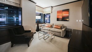 Una sala de estar moderna con un sofá, una mesa de centro y un televisor. Hay una ventana con persianas y vistas a la ciudad.