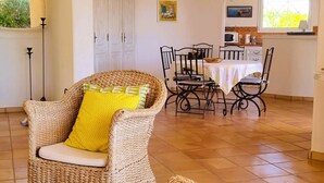 Una silla de mimbre con un cojín amarillo en una habitación con suelo de baldosas, una mesa de comedor con sillas, y una zona de cocina con microondas y armarios.