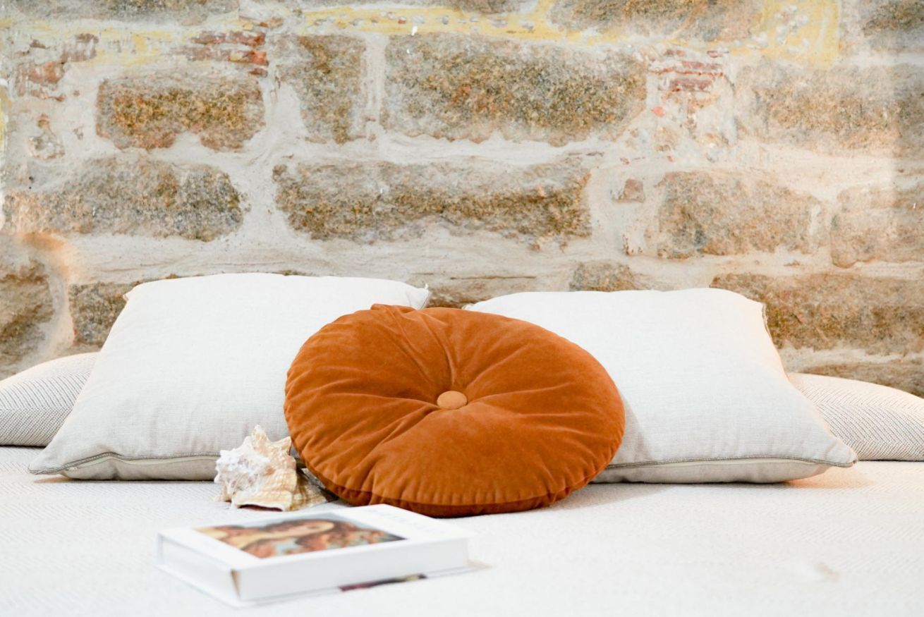 A bed with white pillows and a single orange cushion, a seashell, and a book on a white bedspread against a stone wall.