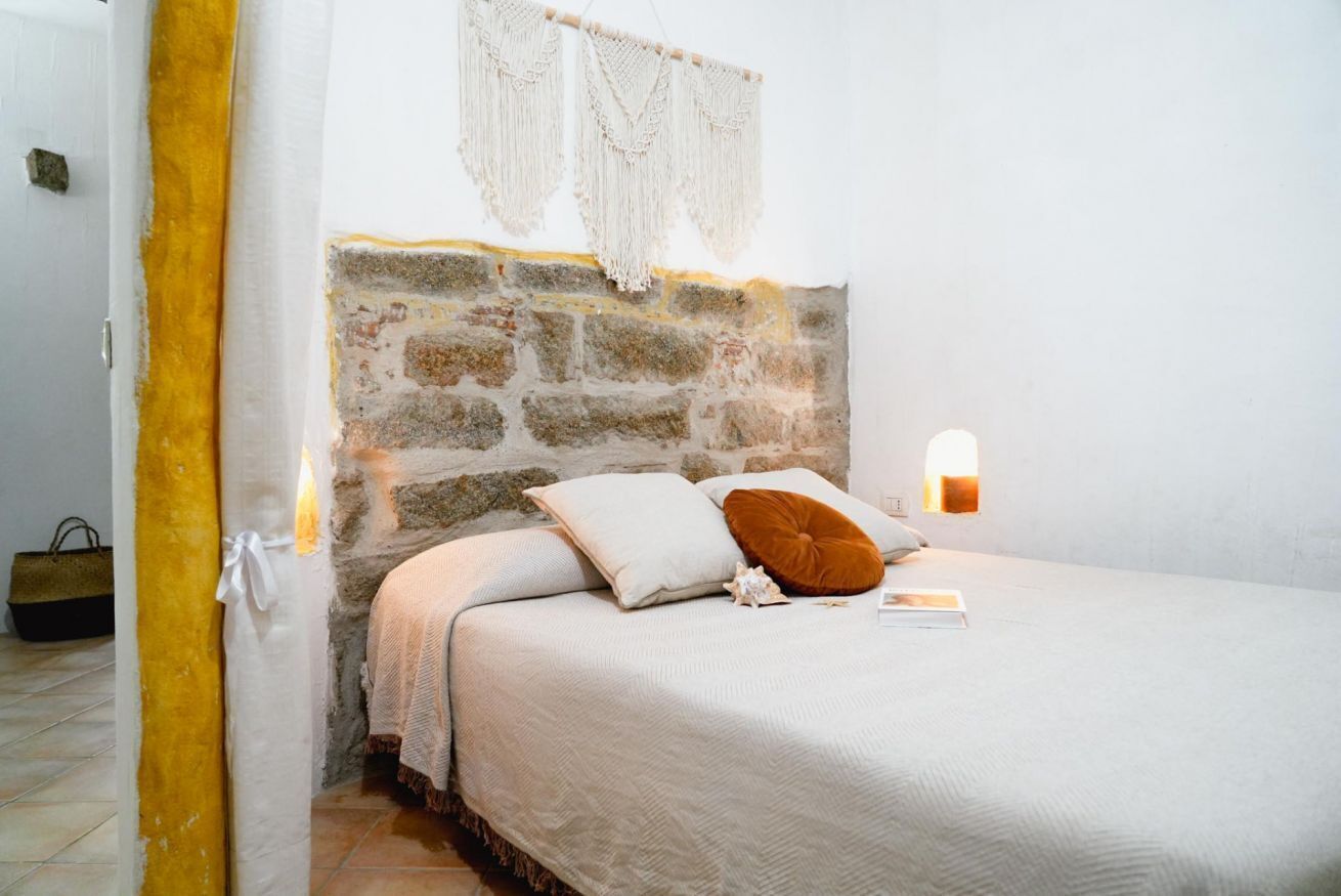 A bedroom with a stone headboard, a bed with white bedding, a brown pillow, and a bedside table with a lamp.