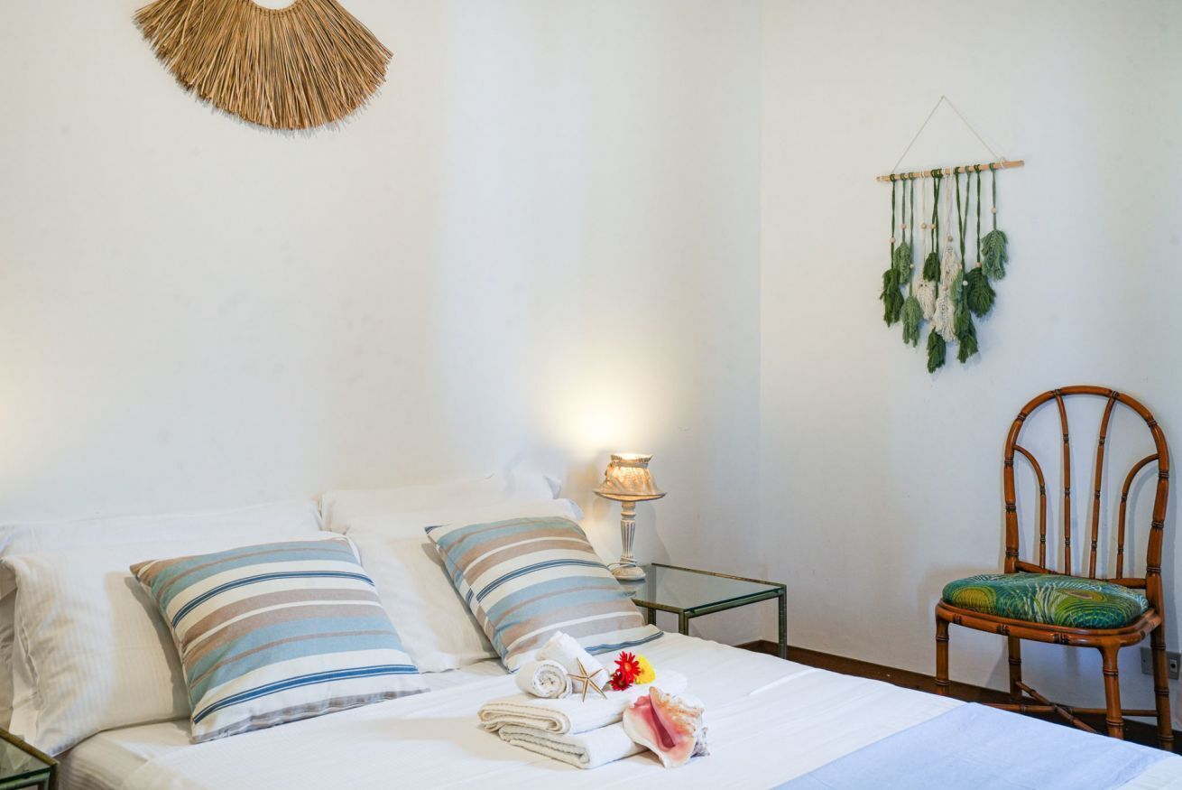A neatly made bed with striped pillows, a small bedside table with a lamp, a woven wall hanging, and a bamboo chair.