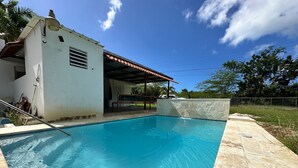 A clear swimming pool with a tiled deck, a covered patio area, and a white building with a red roof. - Sunny Flamboyan Boqueron, 3 bdr/2 bath house w/pool, sleeps 11 (Cabo Rojo)