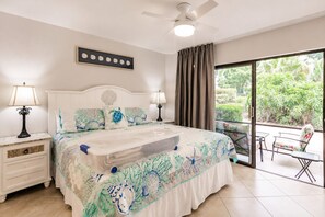 Ein Schlafzimmer mit Bett, Nachttischen, Deckenventilator, Fenster mit Vorhängen und Blick auf Grünfläche.