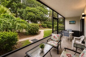 Eine Terrasse mit einem Glastisch und Stühlen, einem kleinen Topfpflanzen und Blick auf einen üppigen Garten mit verschiedenen Bäumen und Sträuchern.