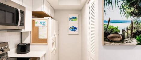 A compact kitchen with a microwave, toaster, and kettle, a corkboard with notes, and a framed tropical beach painting.