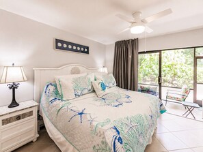 Ein Schlafzimmer mit Bett, Nachttisch, Deckenventilator, einem Fenster mit Vorhängen und Blick ins Grüne.