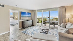 A spacious living room with a large window, a flat-screen TV, a sofa, a coffee table, and a dining area with chairs.